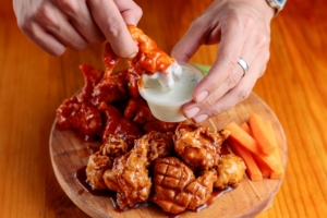 Hands dipping a buffalo chicken wing into a cup of white ranch dressing on a wooden serving board with carrot sticks.