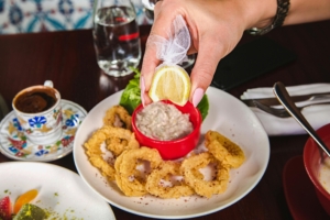 Fried calamari rings served on a white plate with a hand squeezing a lemon wedge over a red dipping sauce.