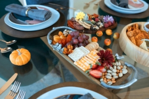 A long wooden charcuterie board filled with crackers, sliced cheese, grapes, strawberries, and nuts on a dining table.