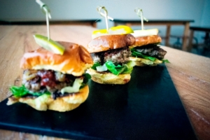 Three beef sliders on mini buns topped with pickles, served on a black slate serving board.