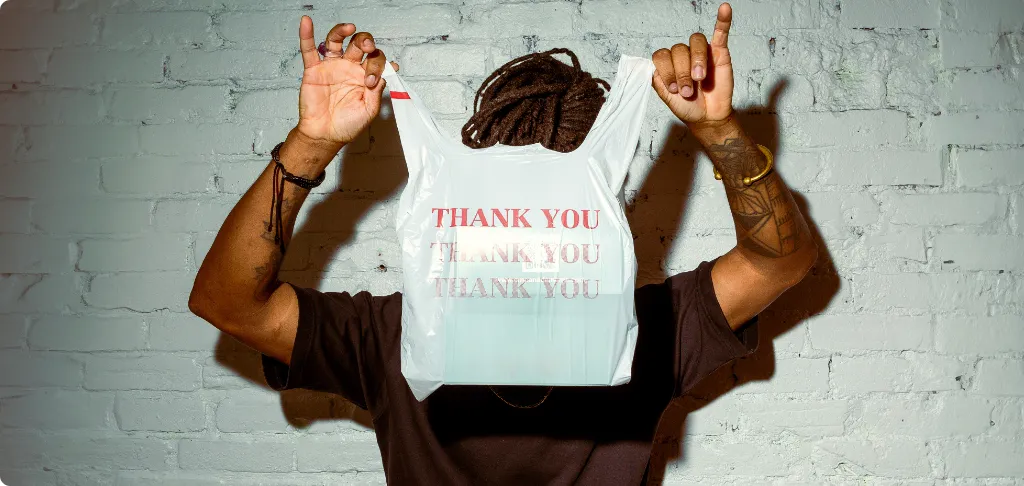 A person holds a white takeout bag reading “THANK YOU” in red, raised above their head against a light brick wall, with arms and tattoos visible and their face mostly hidden by the bag.