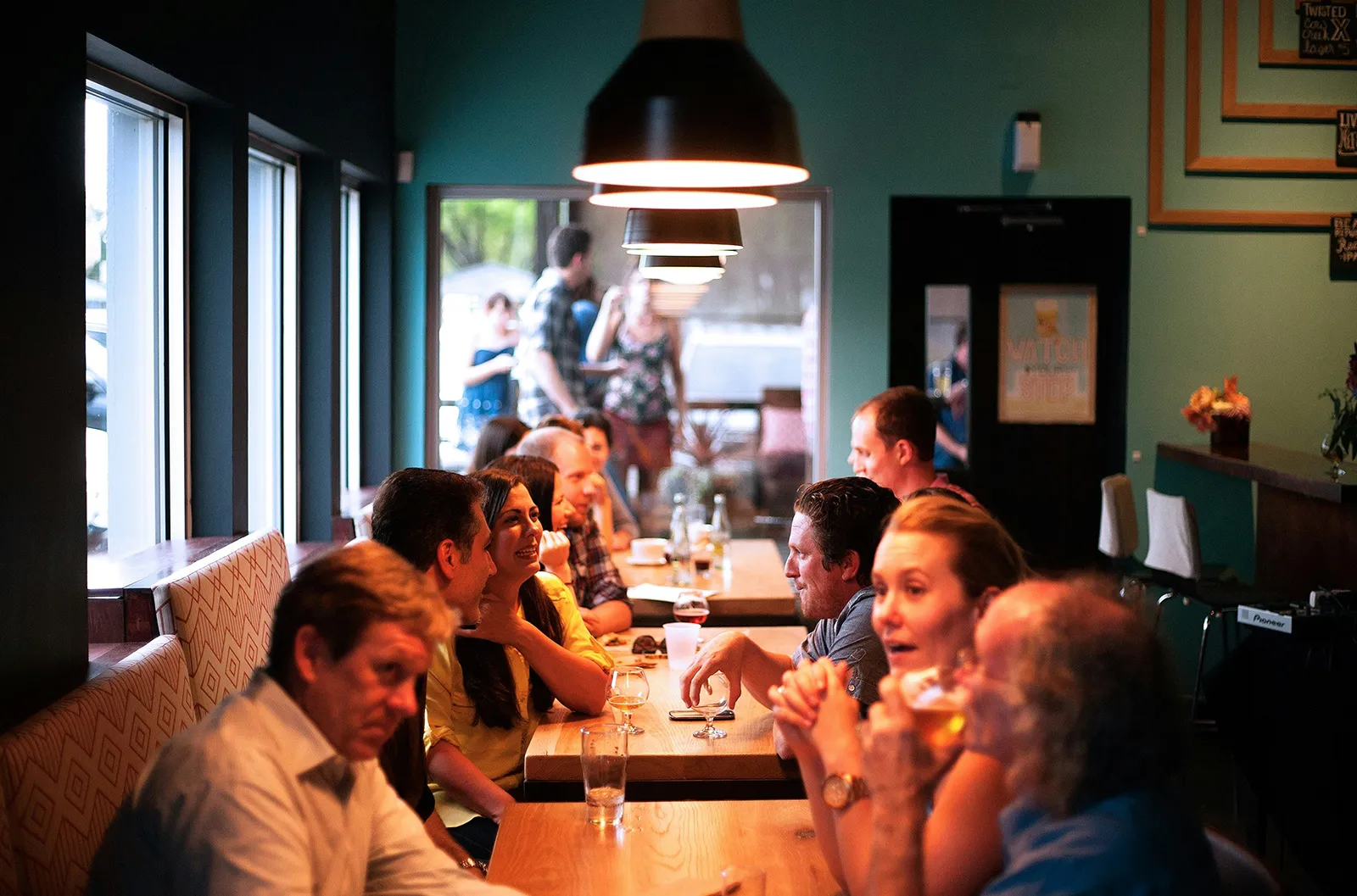 A group of people sitting at a long table in a cozy café or bar, chatting and drinking under warm hanging lights.