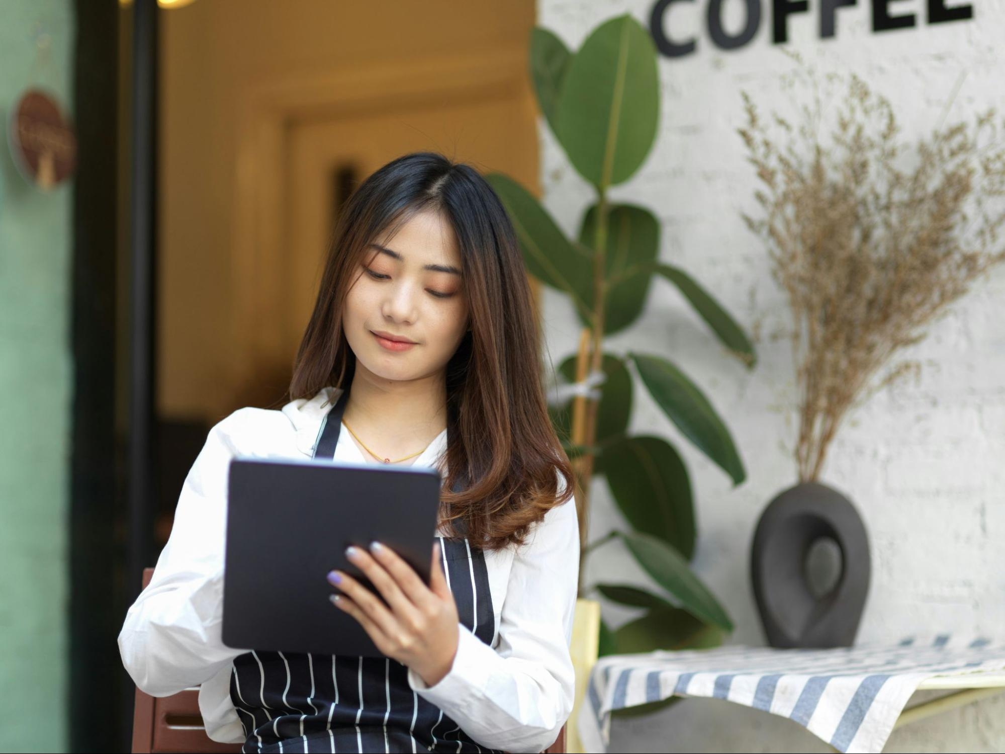 Restaurant staff member in an apron holding a tablet outside a café entrance.