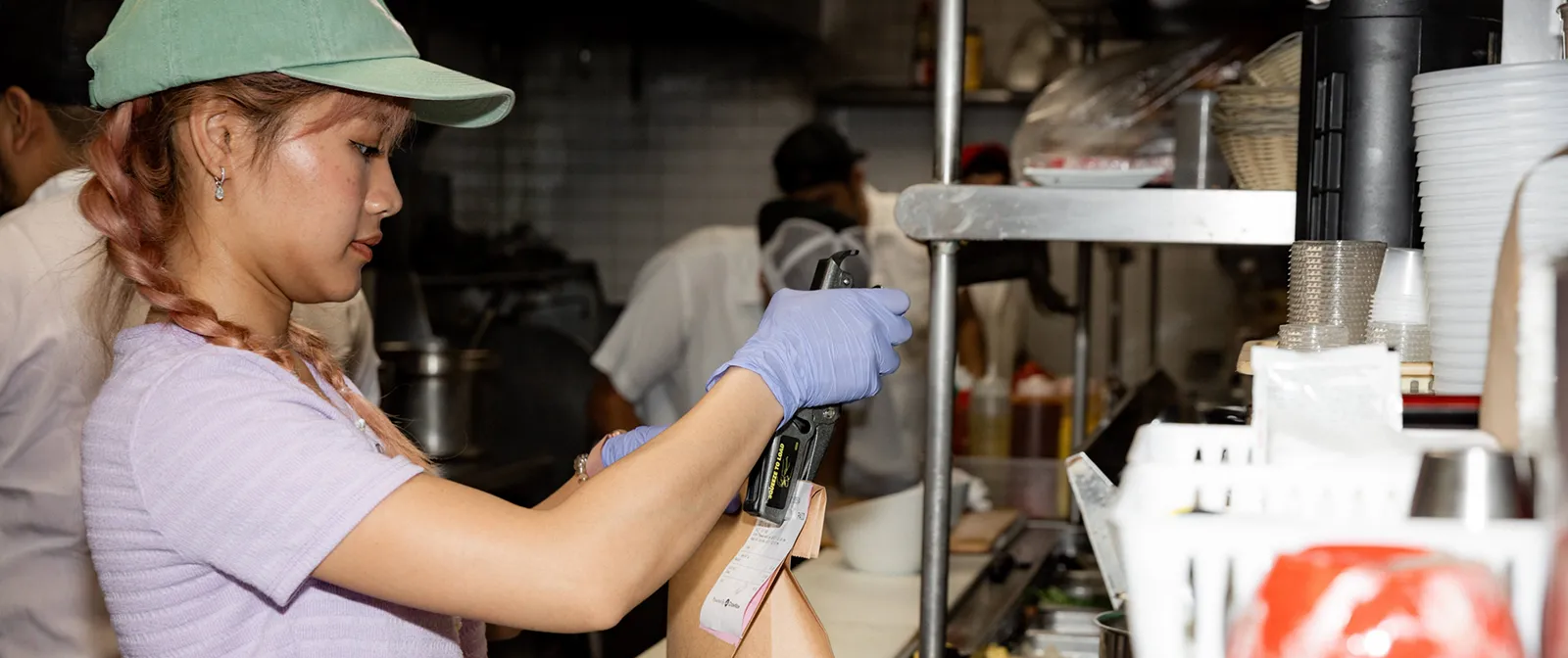 A restaurant worker wearing gloves staples a paper receipt to a takeout bag at a busy kitchen counter, with prepared food and sauces visible in the foreground.