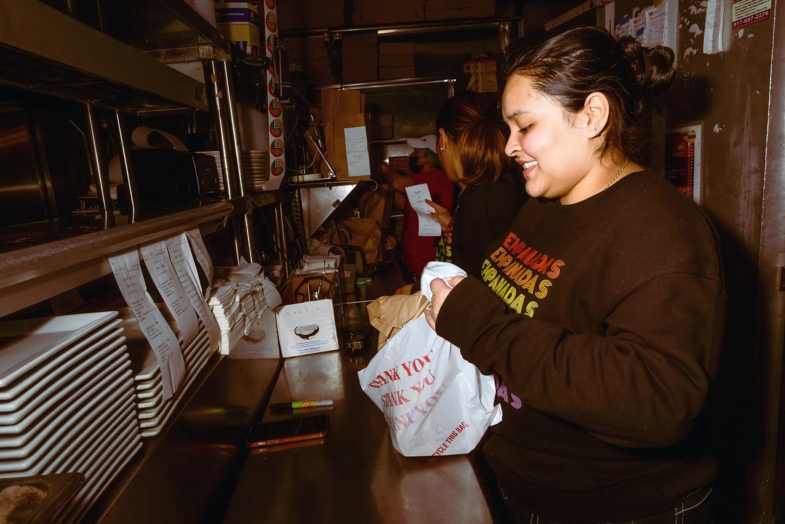 A restaurant worker in a narrow kitchen packing a takeout order, with printed receipts hanging from a rail and stacks of plates and containers on metal shelves.