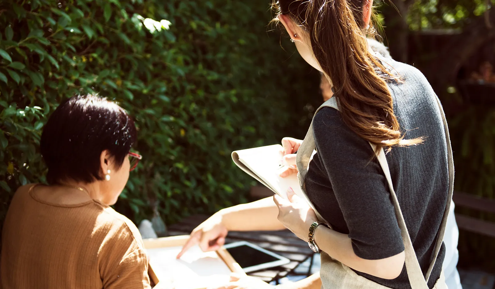 Two women standing outdoors near greenery, placing a food order together as one points to a menu while the other writes selections in a notebook.