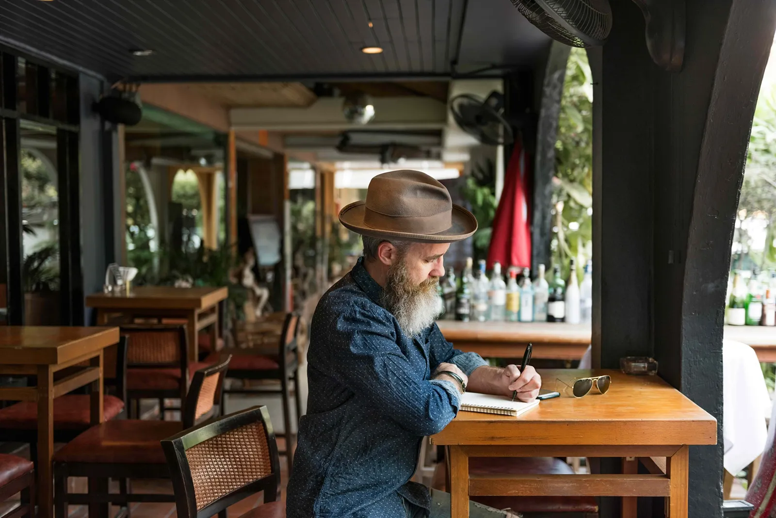 A bearded man wearing a hat sits alone at a wooden café table, writing in a notebook. The setting appears to be an open-air or patio-style café with bottles lined up behind the bar, empty tables around him, and sunlight filtering in, giving the scene a calm, reflective atmosphere.