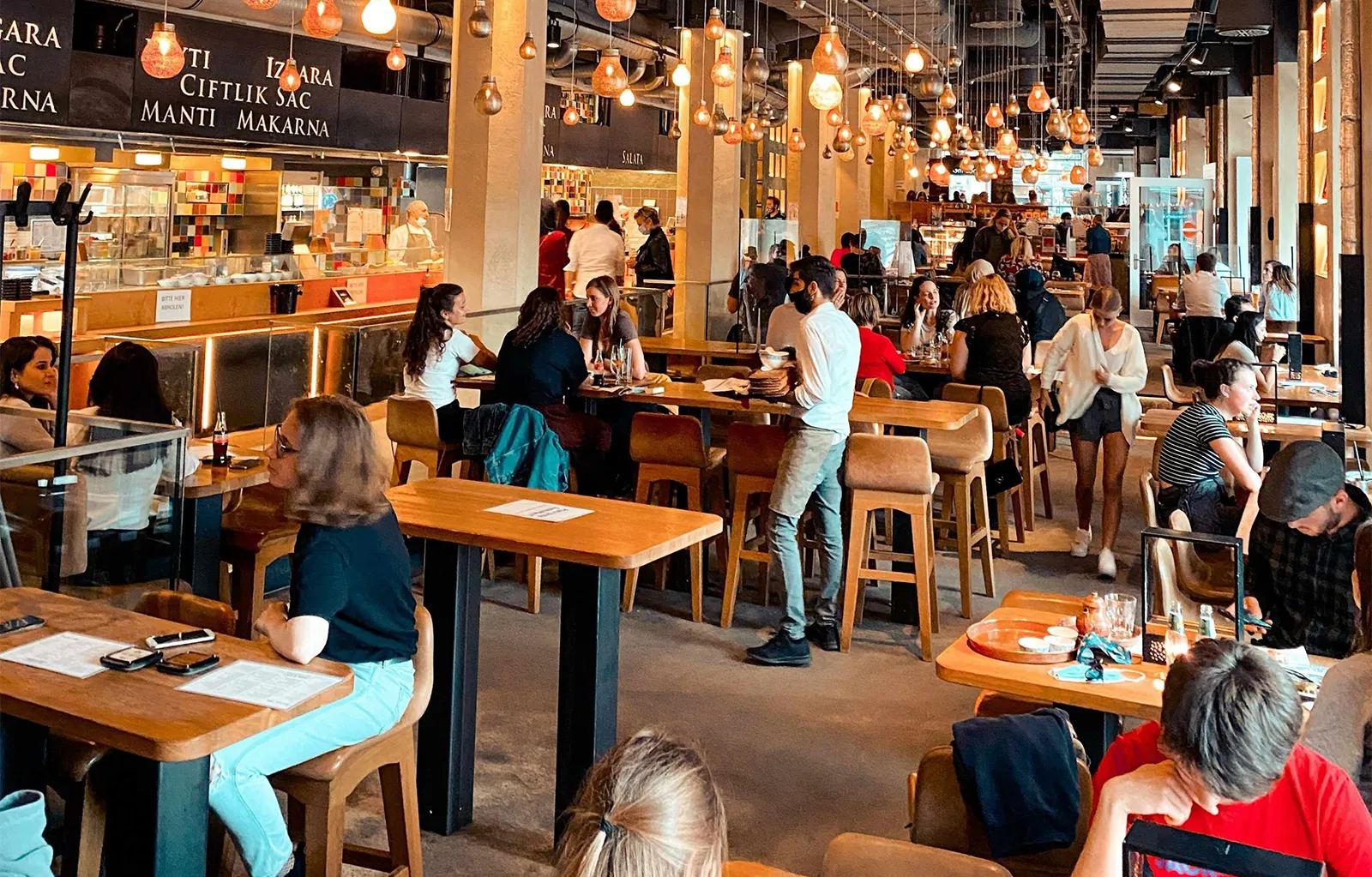 Busy modern food hall with people dining at communal tables, warm hanging lights, and multiple restaurant counters in the background.