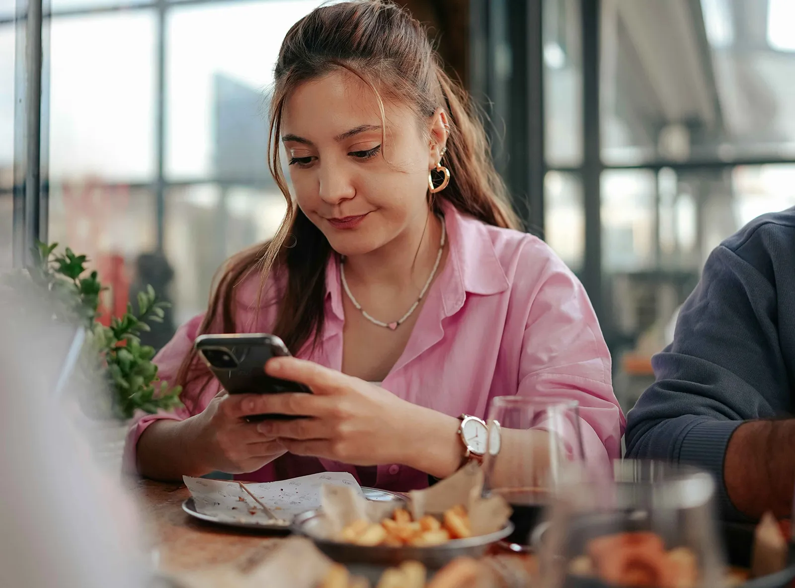 Woman in a pink shirt sitting at a restaurant table, looking at her smartphone with plates of food and drinks in front of her