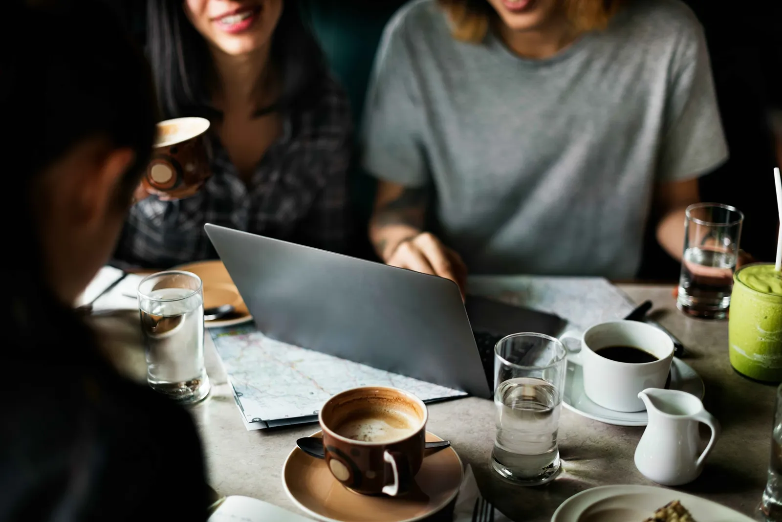 Three people at a café table, smiling and looking at a laptop, with coffee, water, and a green drink on the table—casual work or travel planning vibe.