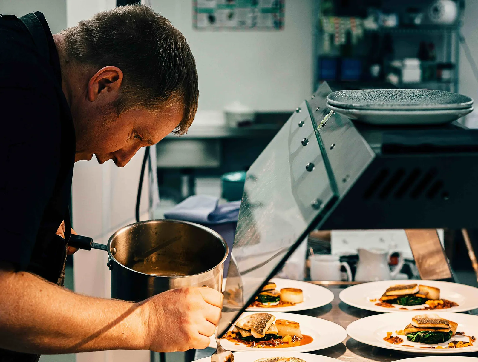 Chef carefully plating sauce onto dishes in a professional kitchen, with multiple plates of prepared fish and vegetables lined up on the counter.