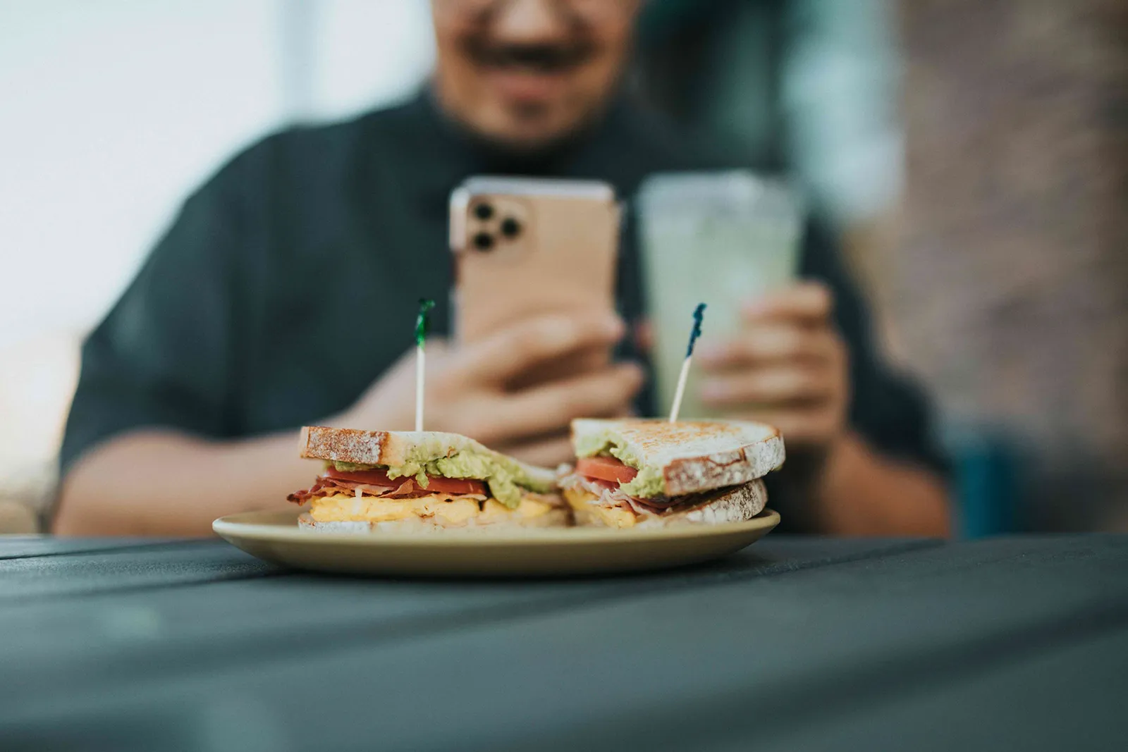 A person photographing a sandwich with their phone while holding a drink.