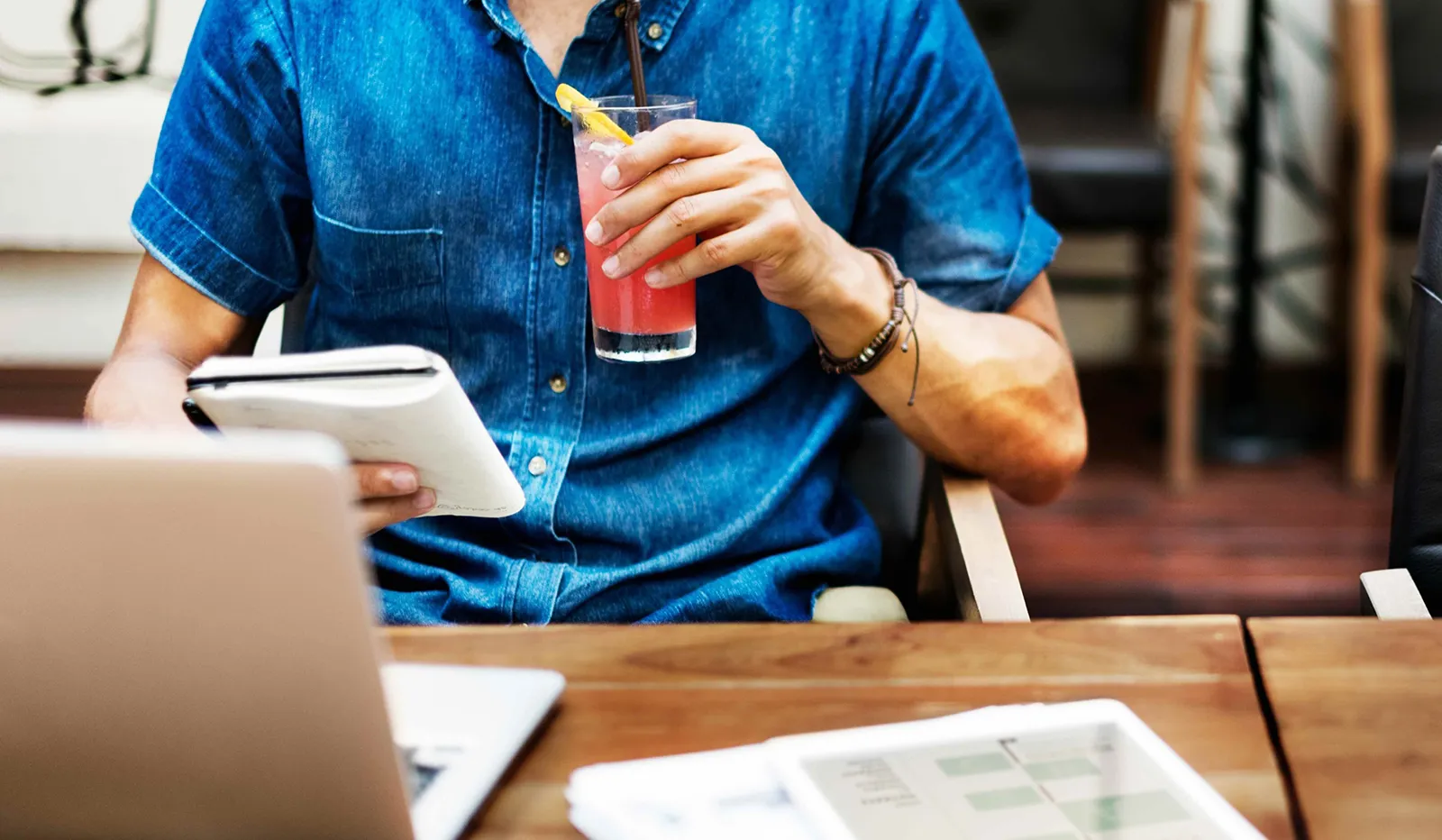A person in a denim shirt sits at a table, holding a pink drink and reading from a notebook, with a laptop and papers in front of them.