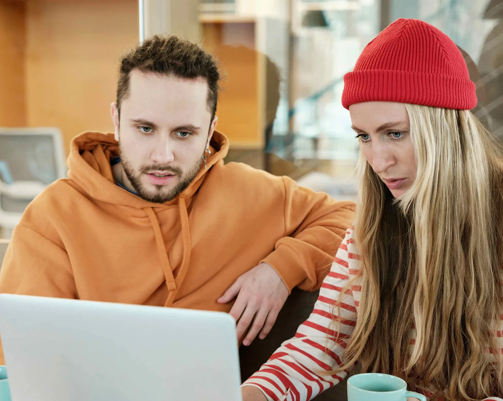 Two people sit side by side in a café, focused on a laptop screen. One wears an orange hoodie and the other a red beanie and striped shirt, with a coffee mug on the table, suggesting they are working or reviewing something together.