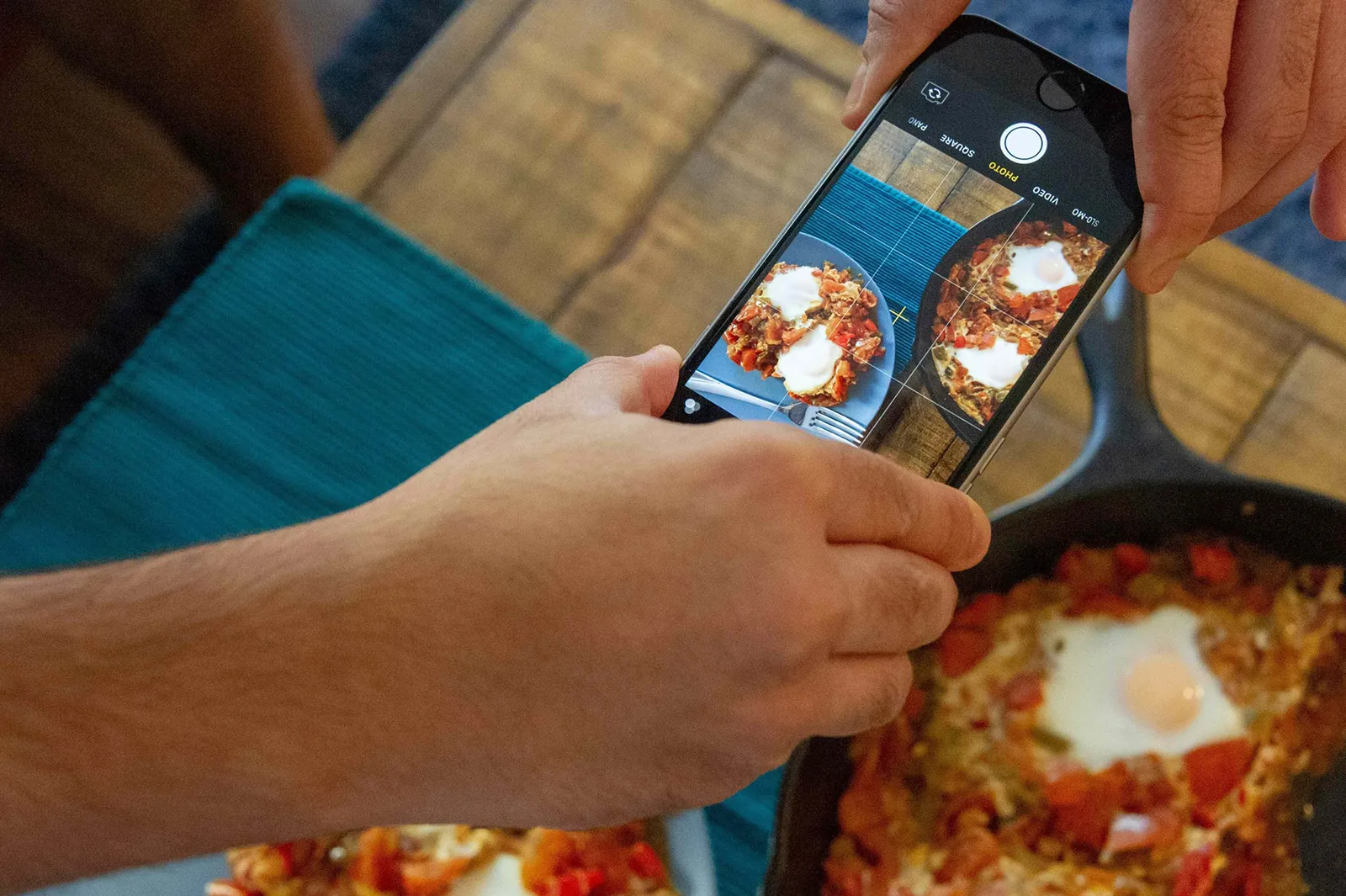 Close-up of hands holding a smartphone to photograph a skillet of food with a sunny-side-up egg, with the camera screen showing the dish being framed for a photo.