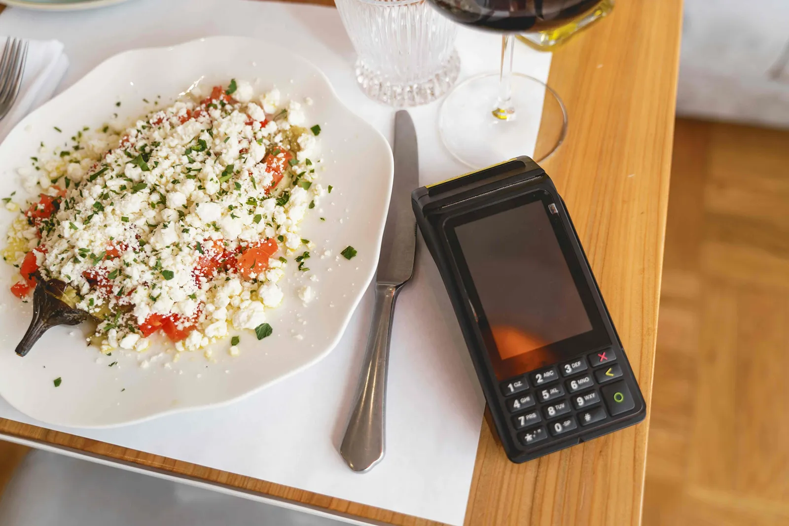 A plate of salad with feta and herbs on a table next to a payment terminal and a glass of wine.