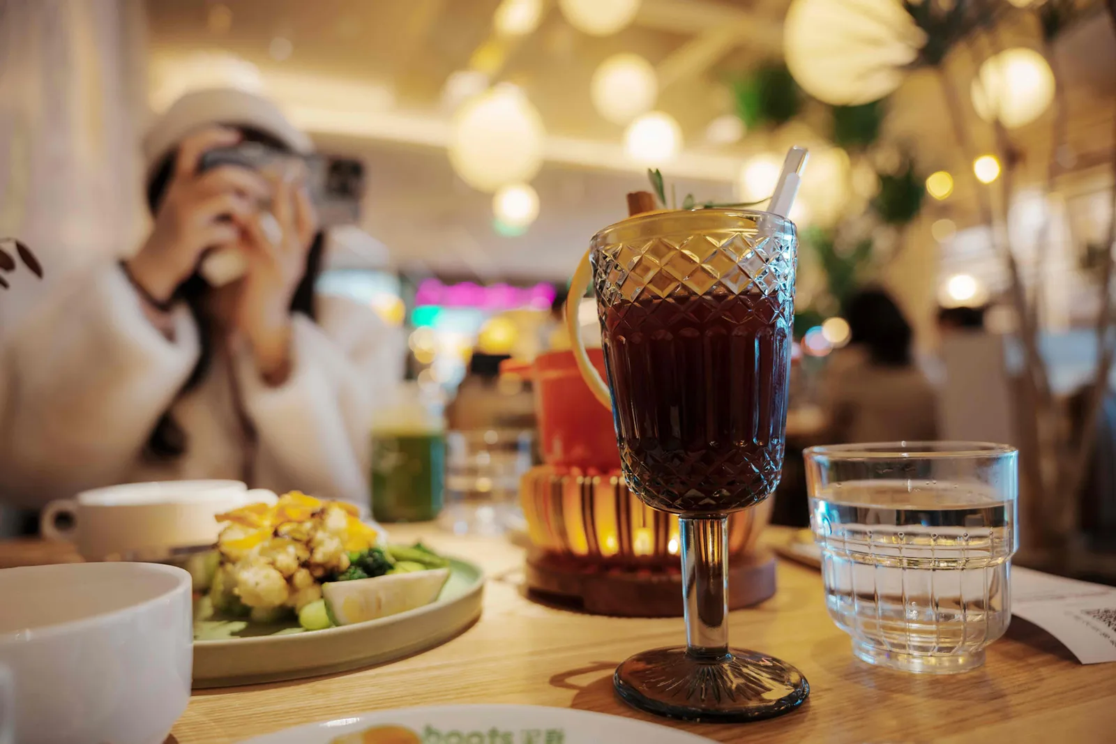A close-up of a cocktail on a café table, with a plate of food and a person in the background taking a photo, under warm, decorative lighting.