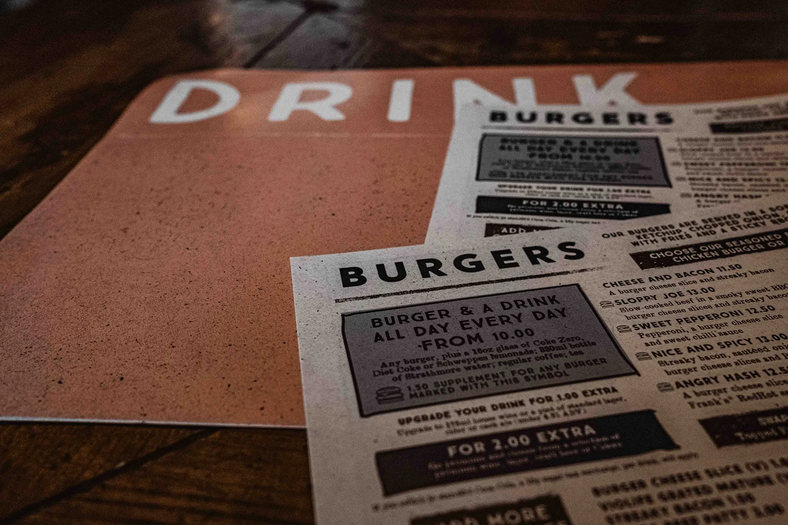 Close-up of a restaurant menu on a wooden table, showing the word ‘DRINK’ on one page and a ‘BURGERS’ section with prices and combo details on the other.