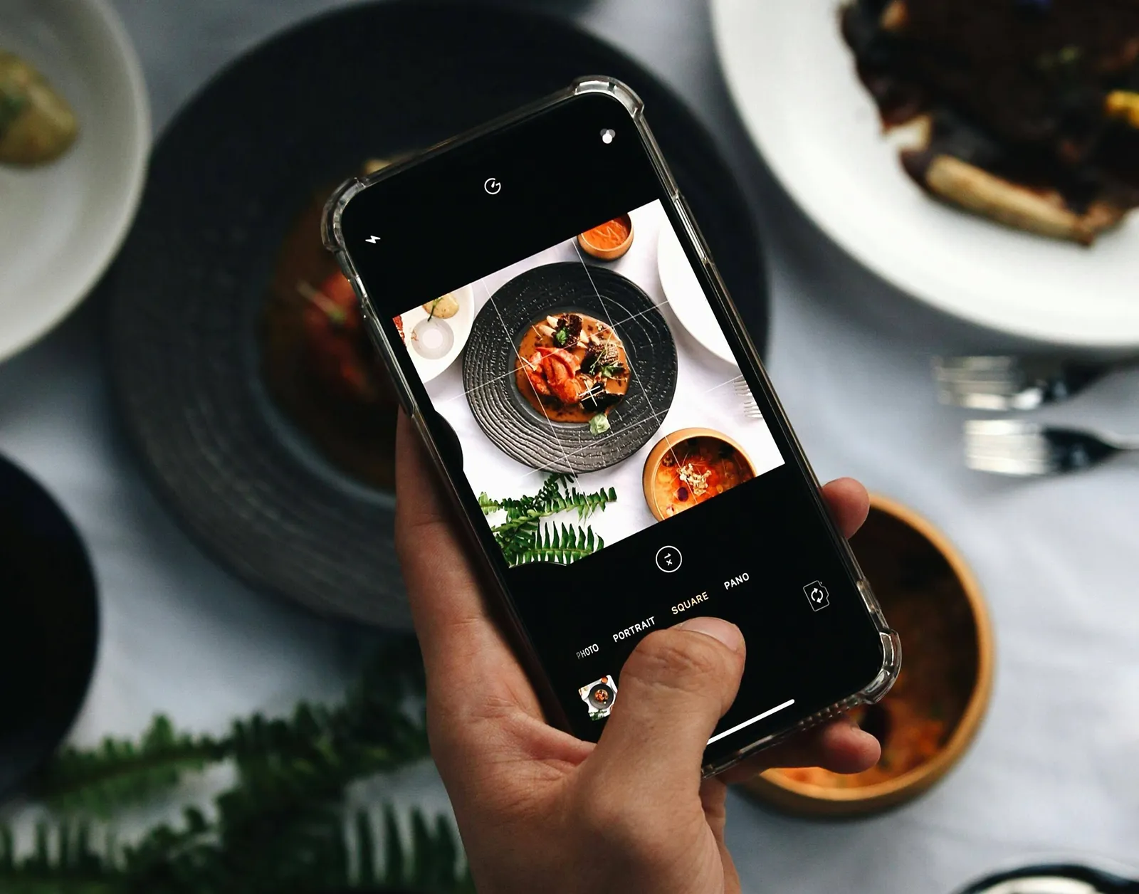 A hand holding a smartphone taking a top-down photo of a plated dish, with other bowls and plates arranged on the table around it.