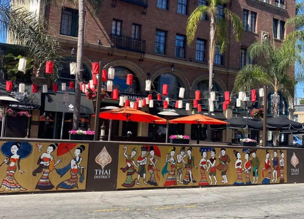 An outdoor café/restaurant frontage with red-and-white lanterns, palm trees, and patio umbrellas, featuring a mural of traditional Thai dancers and the “Thai District” sign along the street barrier.