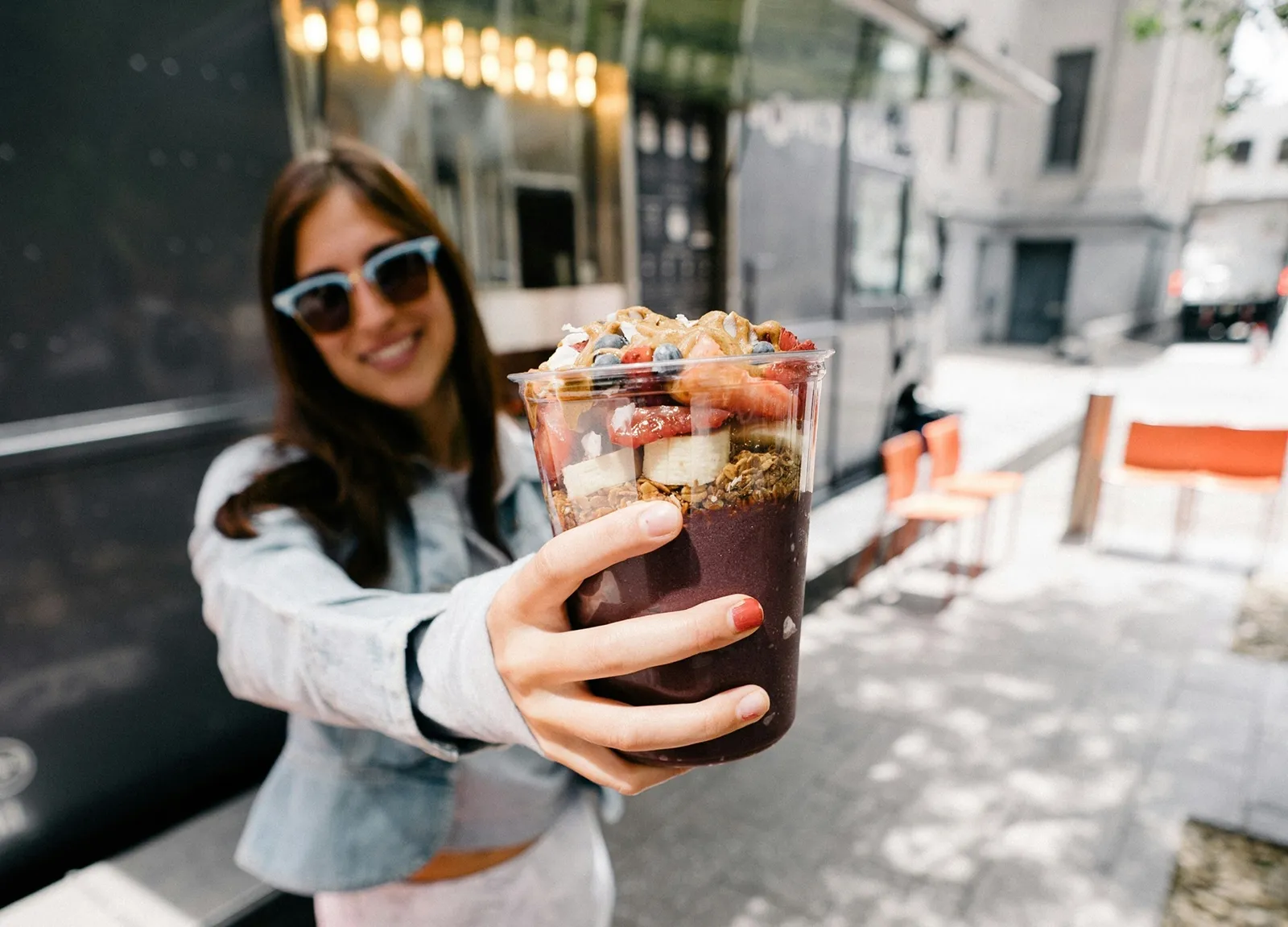 A woman holding out an acai bowl with fruit and granola, likely just picked up from a café or smoothie shop.