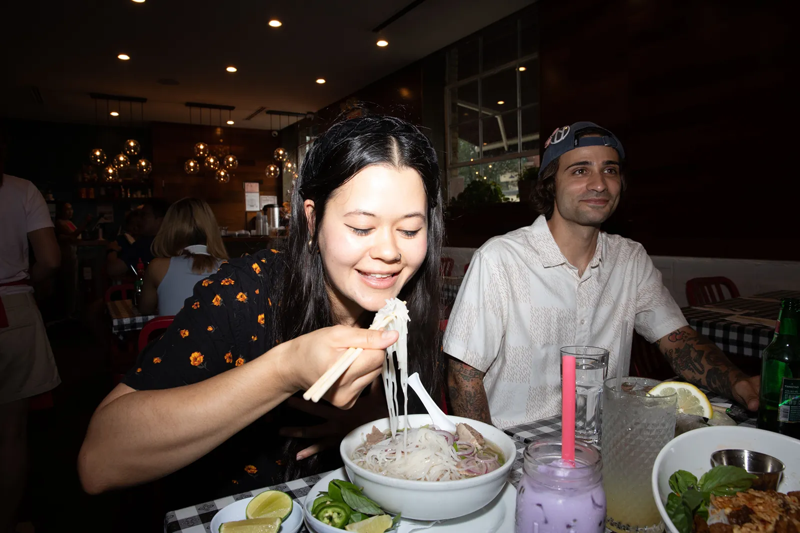 Two people are seated at a restaurant table. The person in the foreground is using chopsticks to lift noodles from a bowl of soup, smiling as they’re about to eat. In front of them are herbs, lime wedges, and drinks, including a purple beverage in a jar and a tall glass with ice and lemon. The person beside them is watching with a relaxed expression, sitting back with their own drink on the table. The setting appears to be a cozy, softly lit café or casual restaurant with other diners and warm pendant lights in the background.