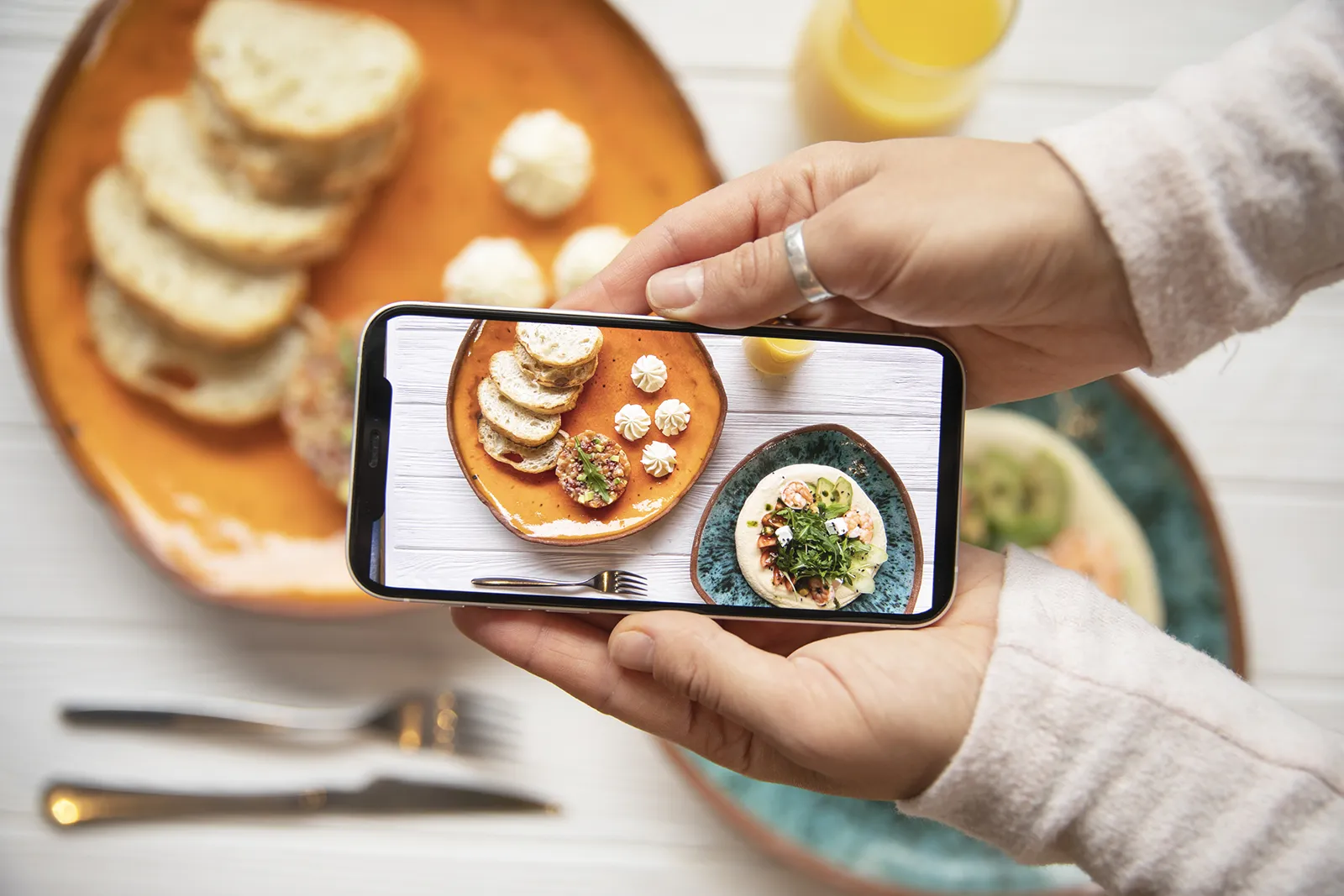 Hands holding a smartphone taking a top-down photo of plated appetizers and toast on a table, with the food framed on the phone screen for social media.