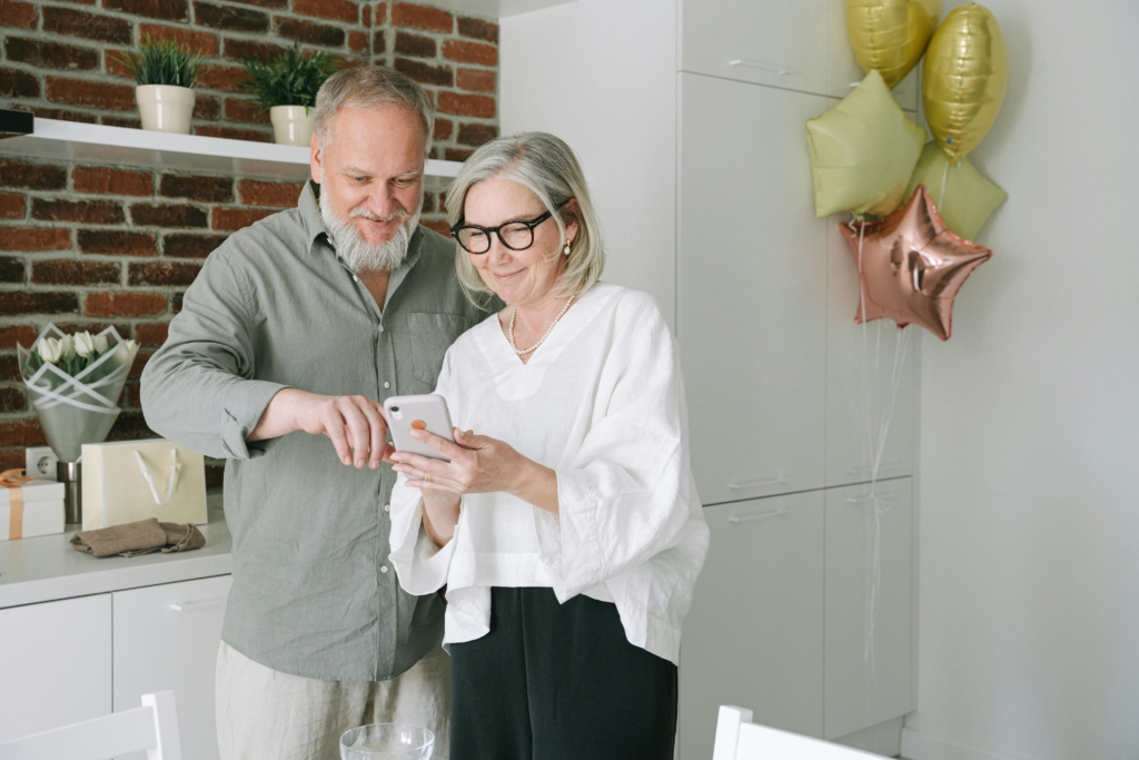 Lifestyle photo of an older couple smiling and using a smartphone together at home, representing accessibility and convenience of voice search technology.