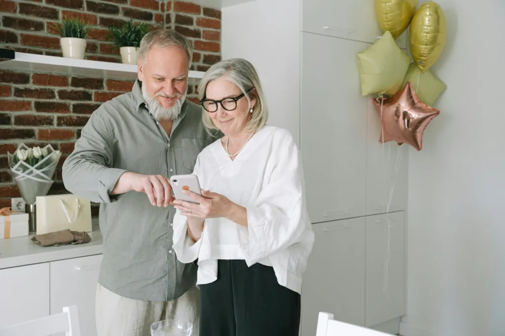 Lifestyle photo of an older couple smiling and using a smartphone together at home, representing accessibility and convenience of voice search technology.