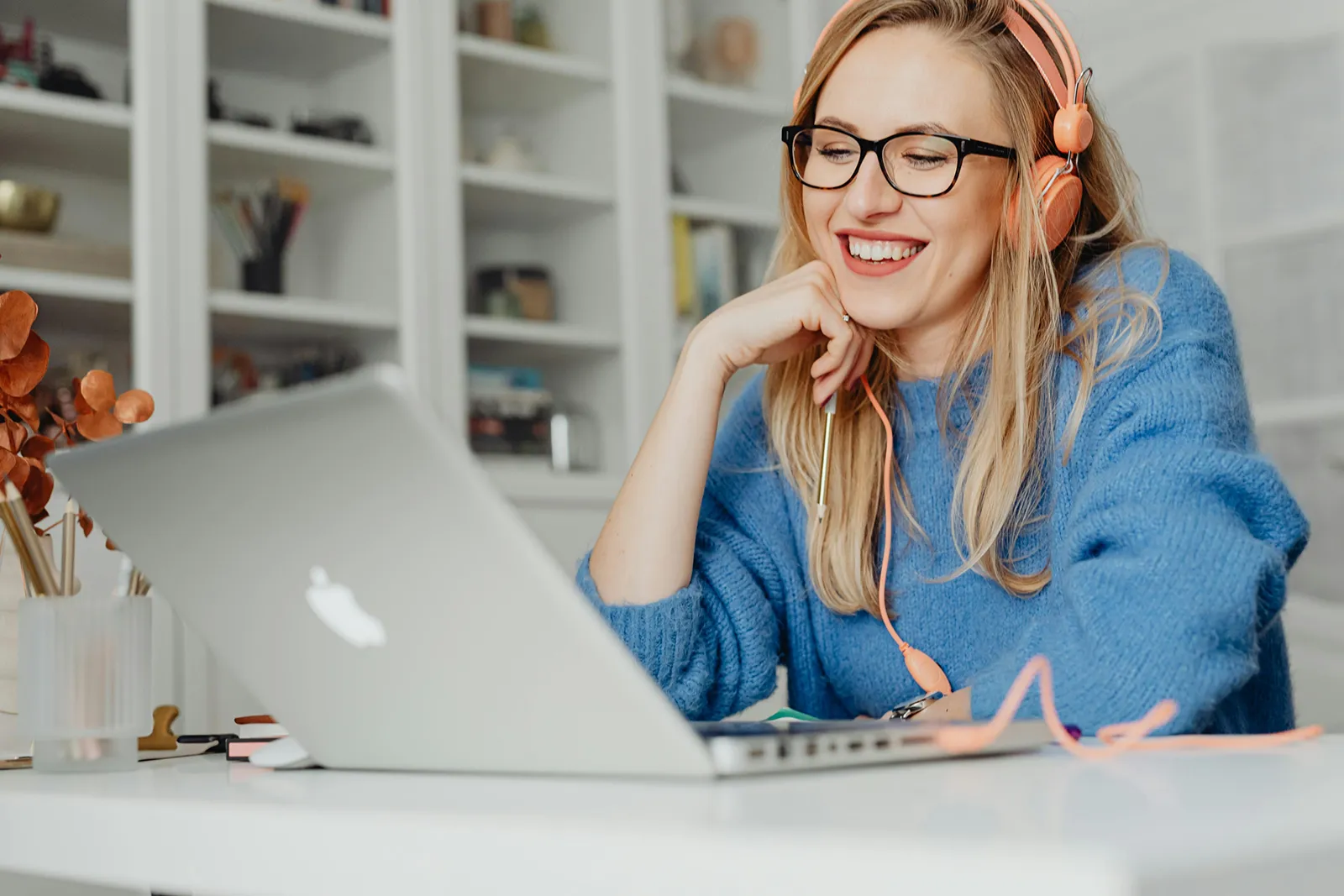 A person is sitting at a desk in a bright, tidy room, smiling while looking at a laptop screen. They’re wearing glasses and over-ear headphones with a cord, and resting their chin on one hand in a relaxed, engaged posture, as if on a video call or listening to something enjoyable. A notebook, pens, and a small decorative plant are on the desk, and shelves with books and objects are visible in the softly lit background, giving the scene a calm, home-office or study-space feel.