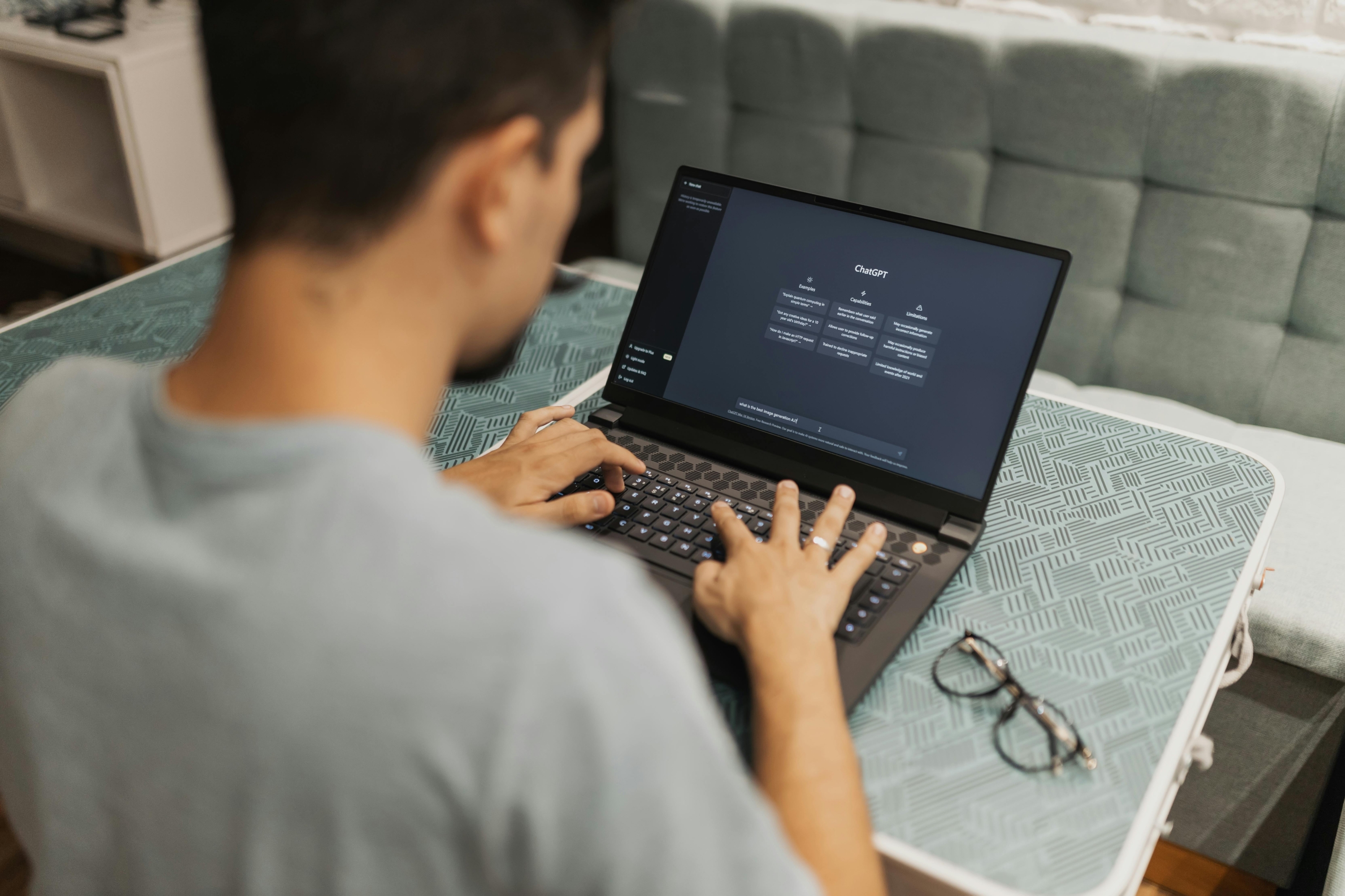photo of a person typing on a laptop with the ChatGPT interface open, sitting at a table with glasses beside the computer