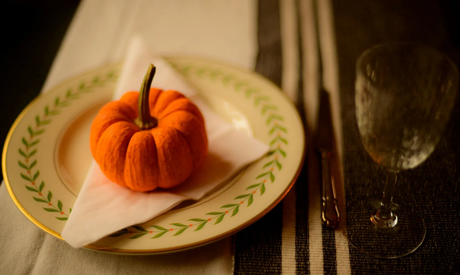 A small pumpkin placed on a decorative plate with a folded napkin, set on a dining table with glassware and cutlery.