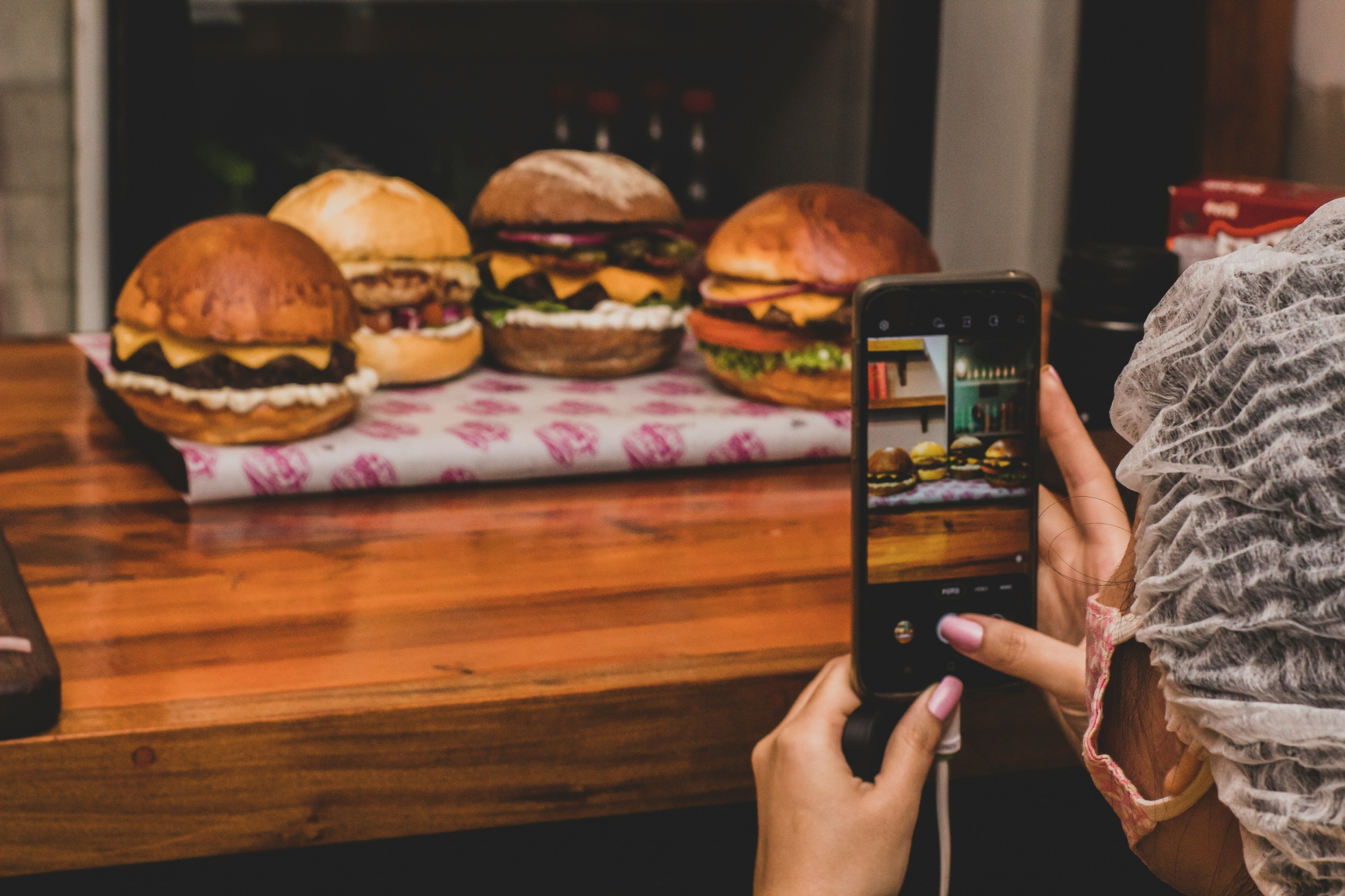 Lifestyle photo of a person taking a smartphone photo of four burgers lined up on a counter, highlighting behind-the-scenes restaurant photography.