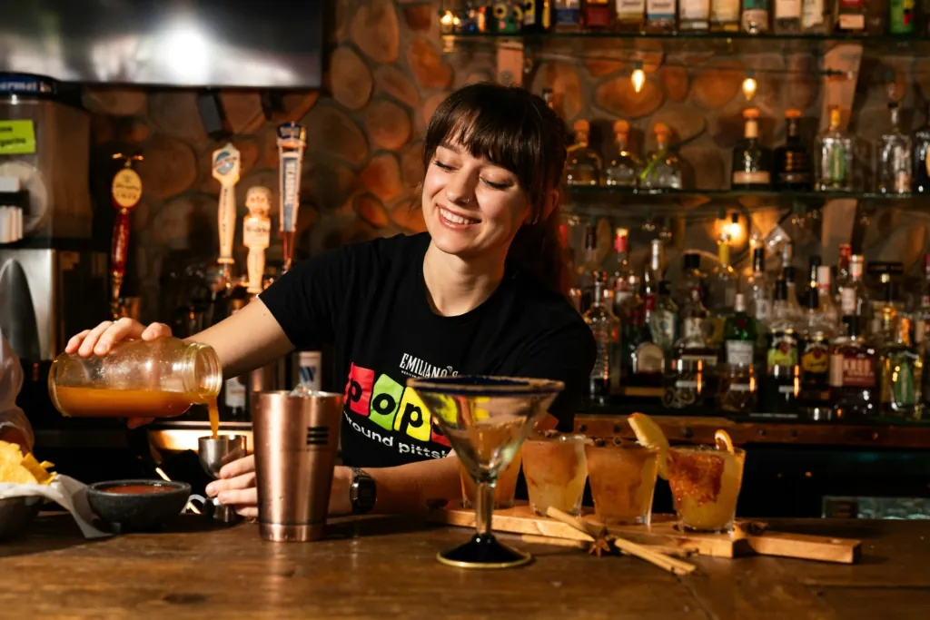 A photo of a smiling bartender pouring a drink from a jar into a shaker, with cocktails and garnishes on the bar.