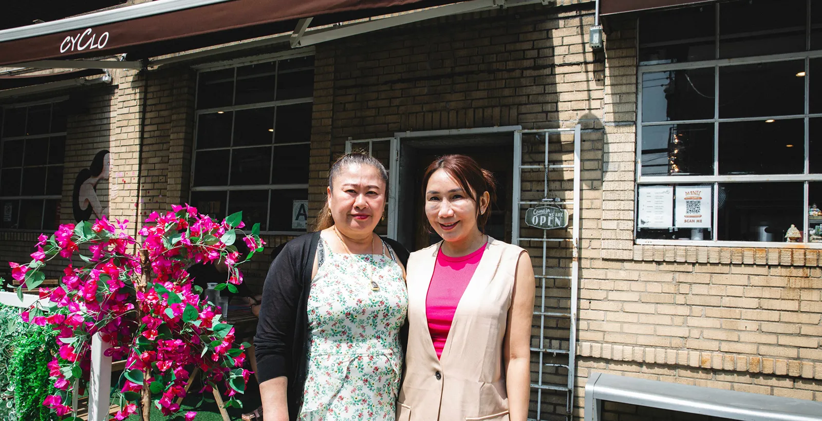 Two women standing outside a brick-front restaurant with an ‘Open’ sign and pink flowers, smiling at the camera in front of the entrance.