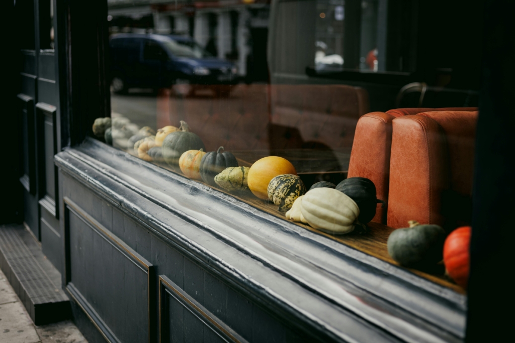 A photo of assorted gourds and pumpkins lined up in a restaurant window display with orange seating inside.