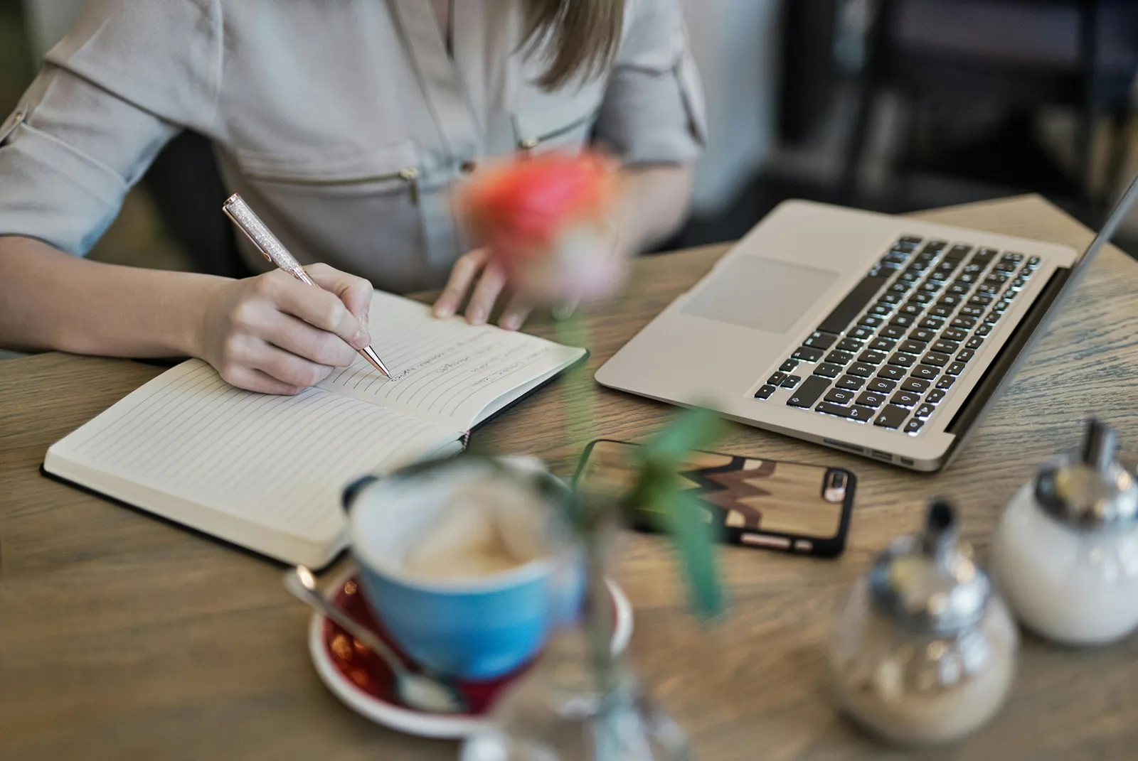 A person sits at a café table, writing in a notebook with a pen next to an open laptop. A cup of coffee, a smartphone, and small table items are visible in the foreground, suggesting a work or study session in a cozy café setting.
