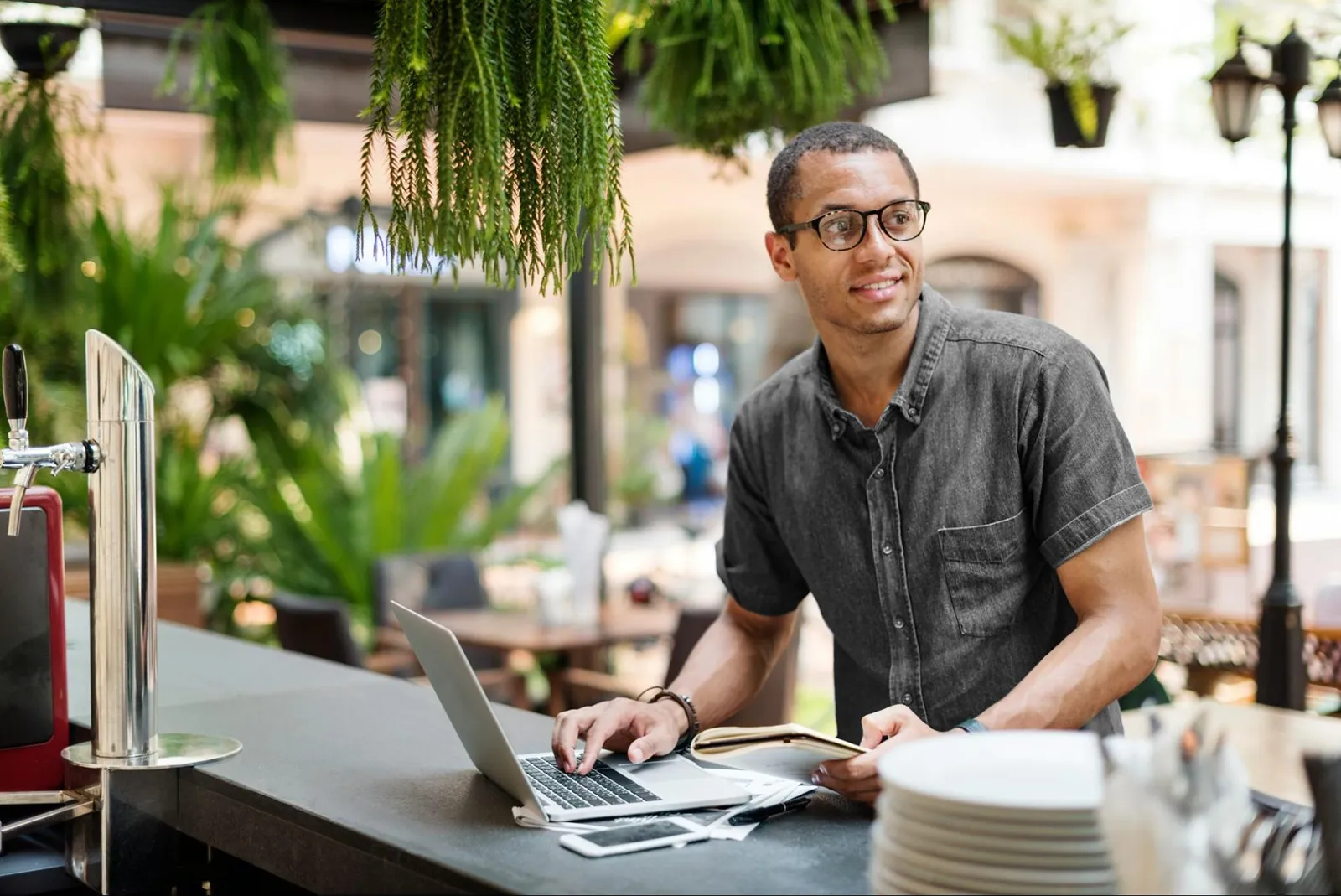 A person working on a laptop behind a café counter, holding a small notebook, in a bright, plant-filled space.