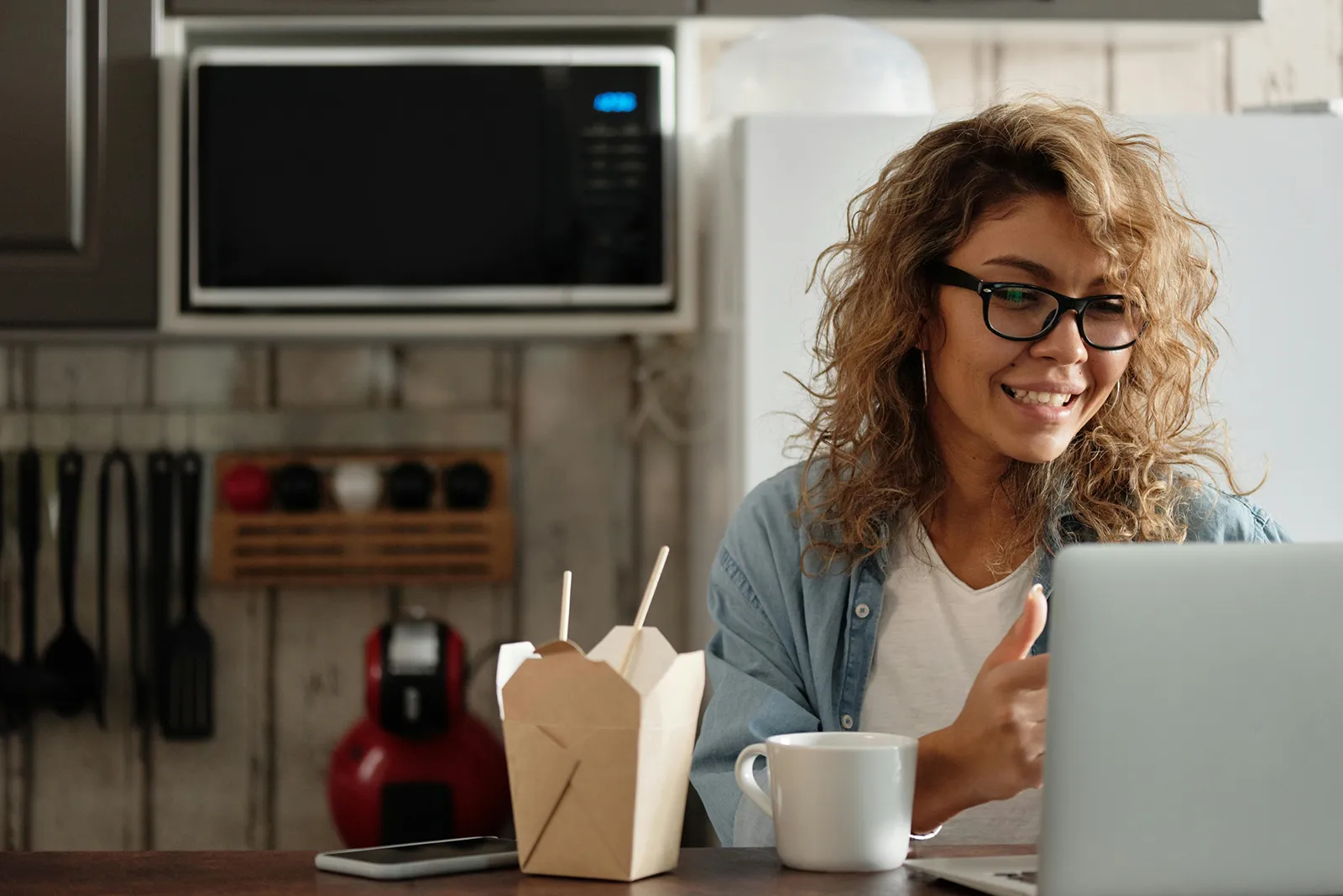 Smiling woman wearing glasses sits at a kitchen table with a laptop, a coffee mug, and a takeout box, appearing to place or review an online food order.
