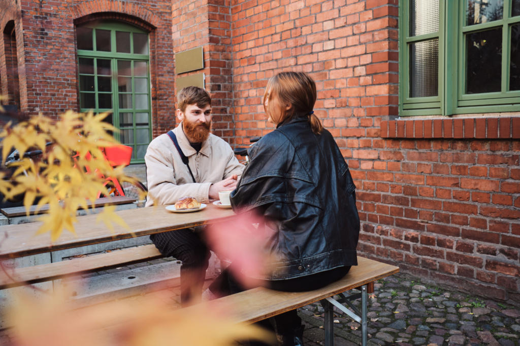 A photo of a couple sitting at an outdoor café table by a brick building, enjoying coffee and pastries in fall weather.