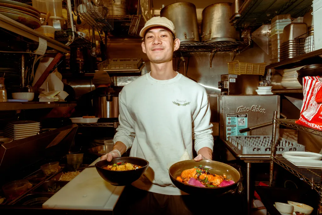 Food runner holding two bowls of plated dishes while standing in the kitchen.