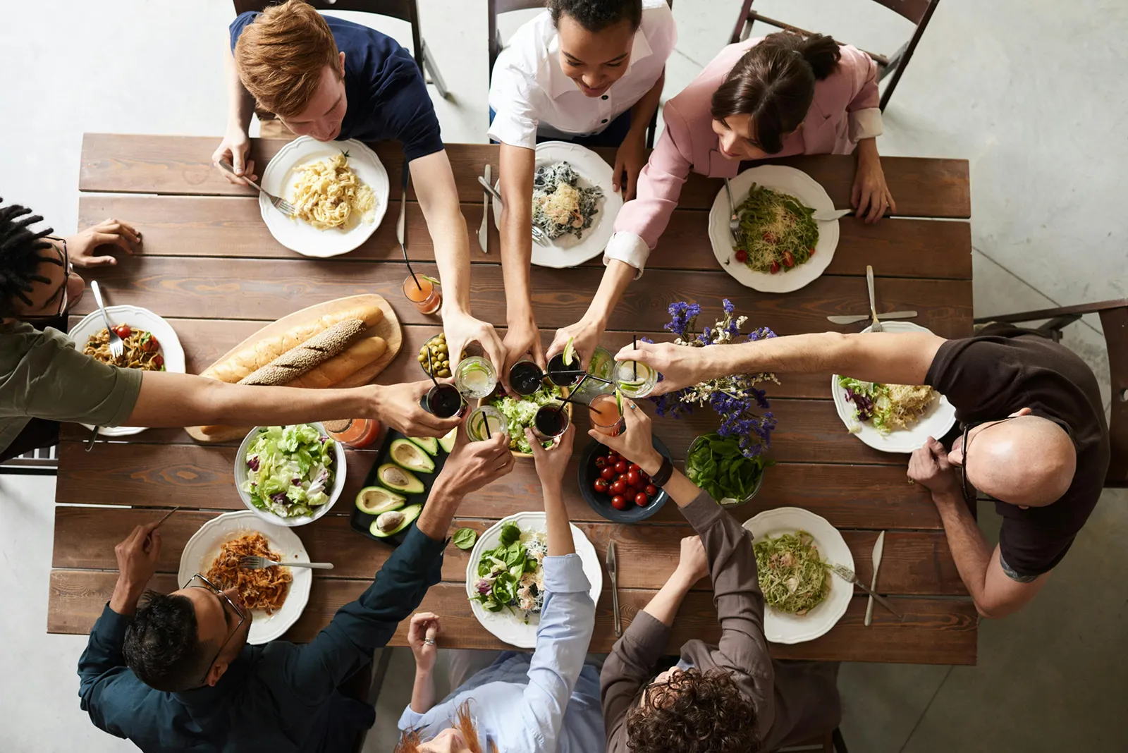 Overhead view of a group of people seated around a wooden table, clinking glasses in a toast over plates of pasta, salad, bread, and drinks.