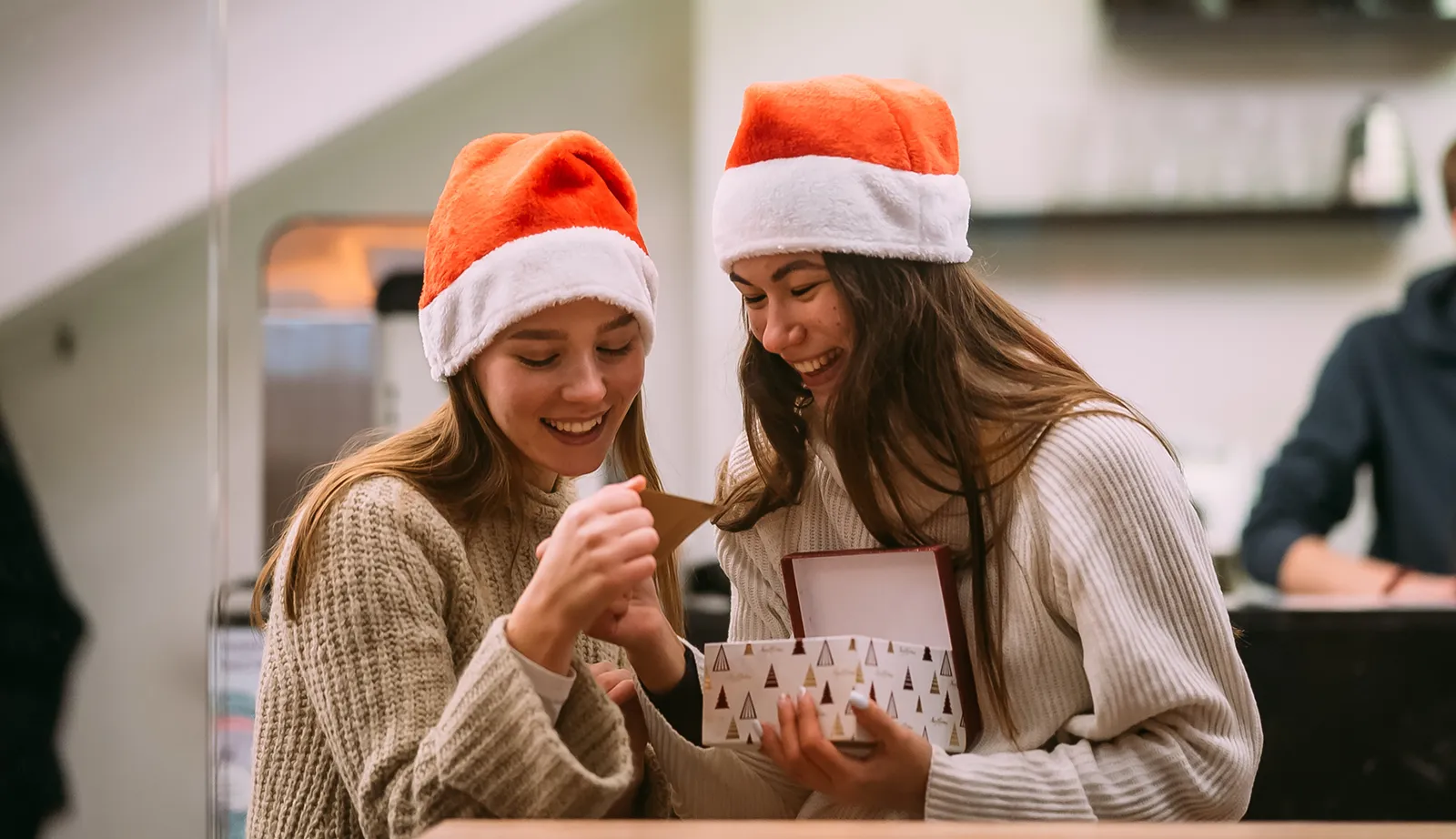 Two young women wearing Santa hats smile as they open a small gift box together, standing in a cozy indoor setting with shelves and people blurred in the background.