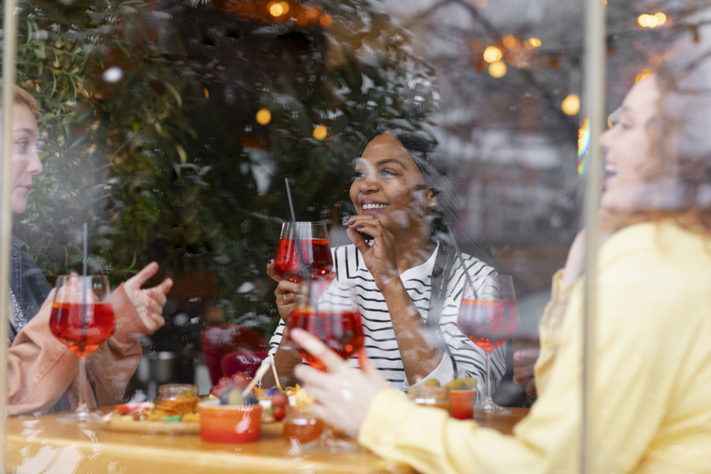 A photo of friends enjoying drinks and appetizers at a restaurant, seen through a window with reflections and festive lights.