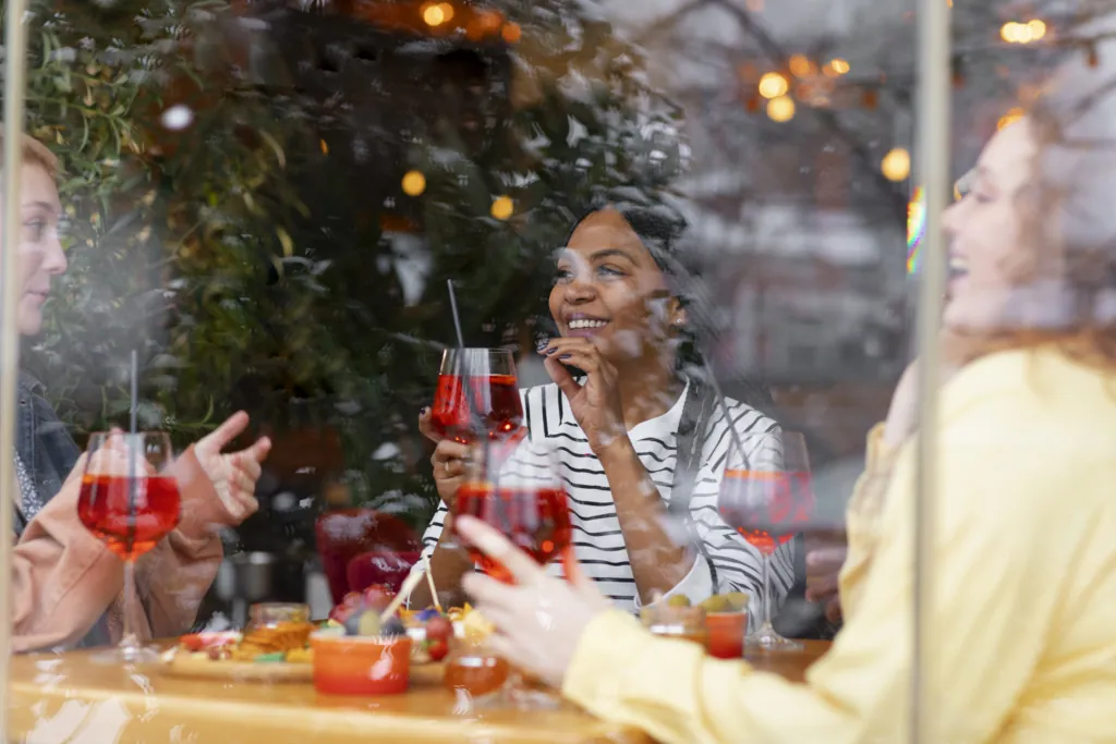 A photo of friends enjoying drinks and appetizers at a restaurant, seen through a window with reflections and festive lights.
