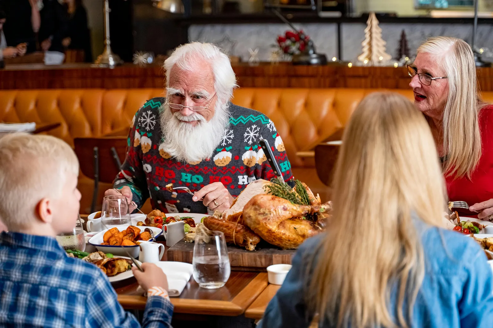 An older man with a white beard in a festive sweater sits at a restaurant table with family, smiling as a roast turkey and side dishes are served, while two children and an older woman look on.
