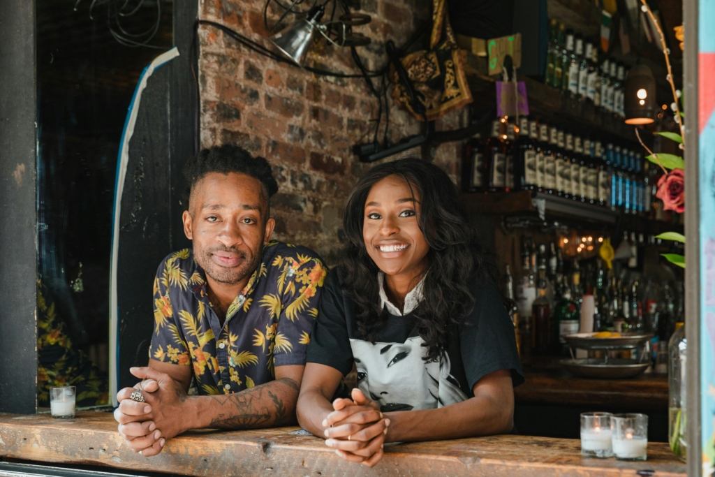 photo of two restaurant owners smiling behind a rustic brick bar.