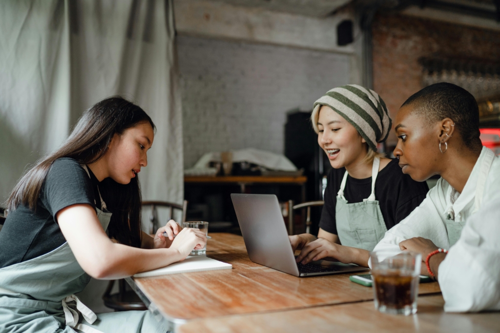A photo of three restaurant staff members in aprons sitting at a wooden table, reviewing notes and a laptop during an operations check-in.