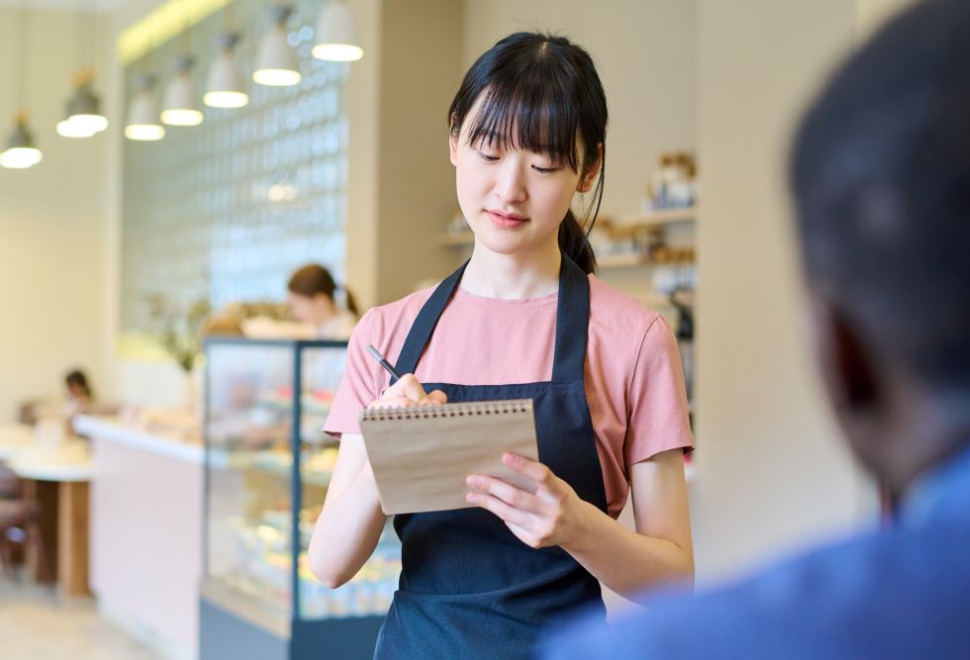 A photo of a restaurant server in a black apron writing an order on a notepad while standing in a bright café.