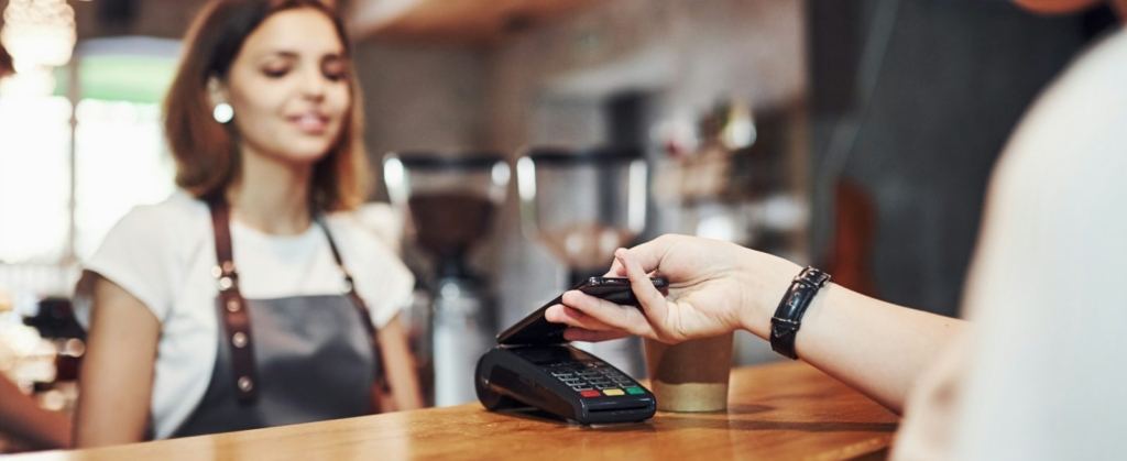 A photo of a customer using a smartphone for contactless payment at a café counter while the barista smiles behind the register.